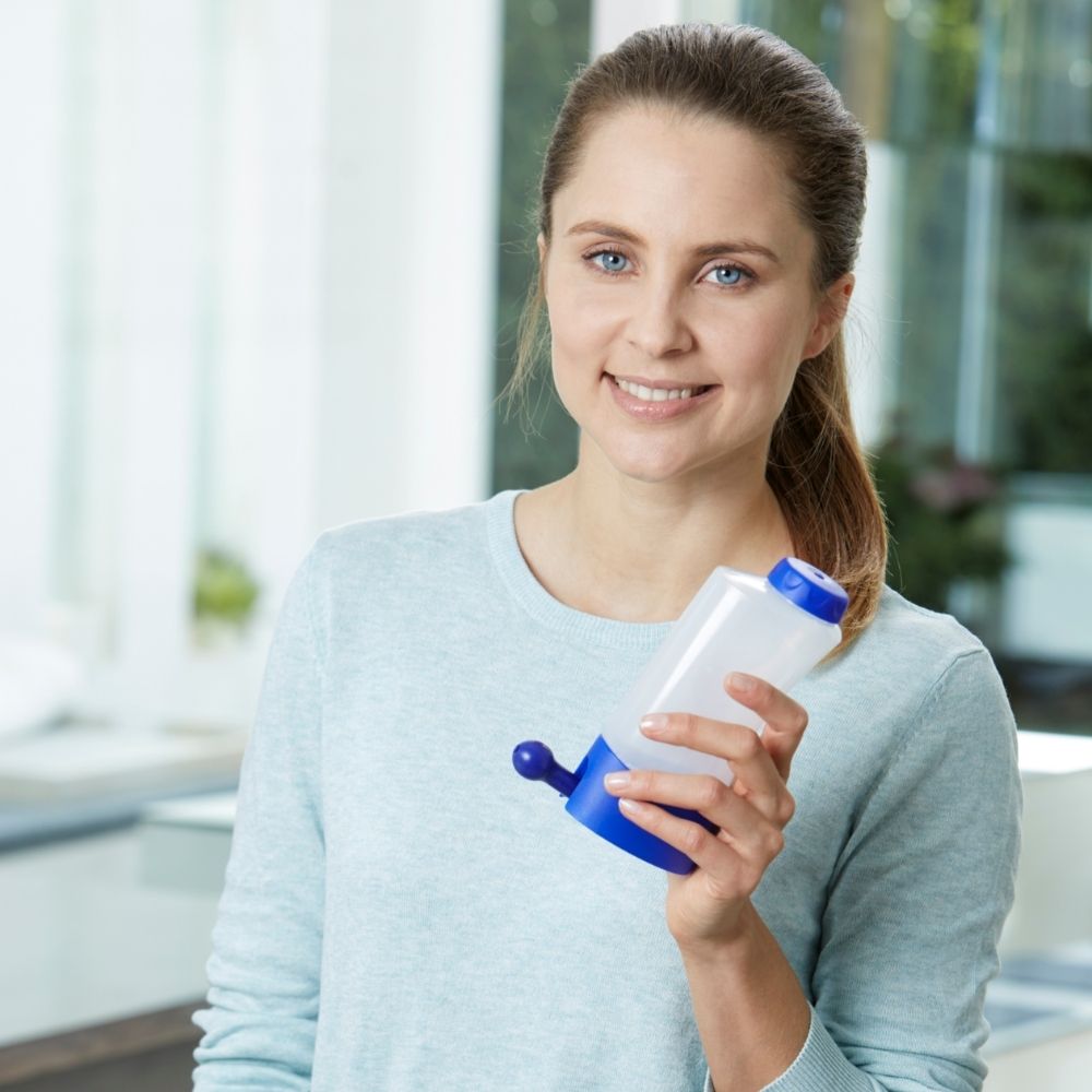 Een vrouw houdt een neusdouche vast met een blauwe dop en basis. Ze lacht in de camera.