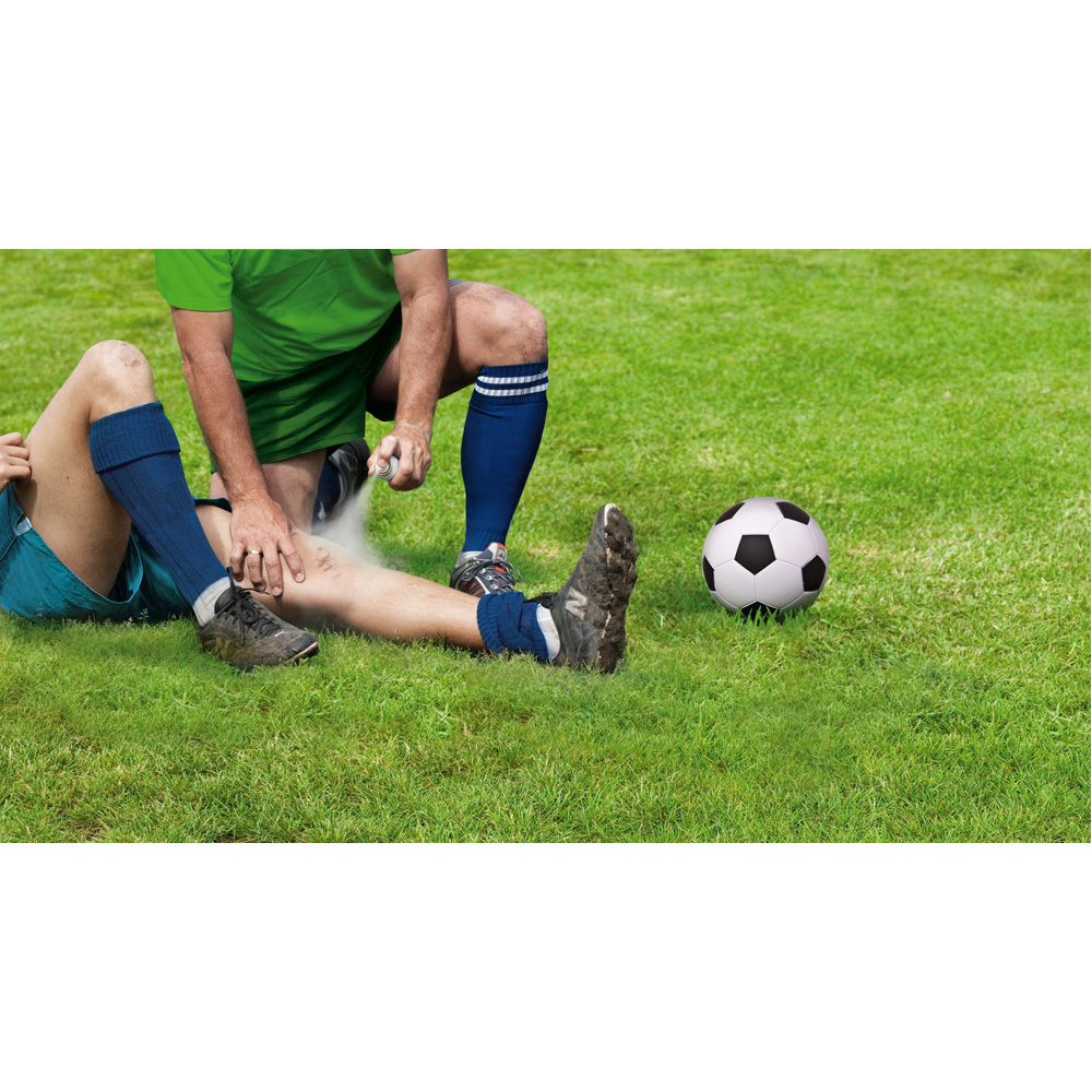 Twee personen op gras. Eén persoon spuit op het been van een ander. Voetbal ernaast.