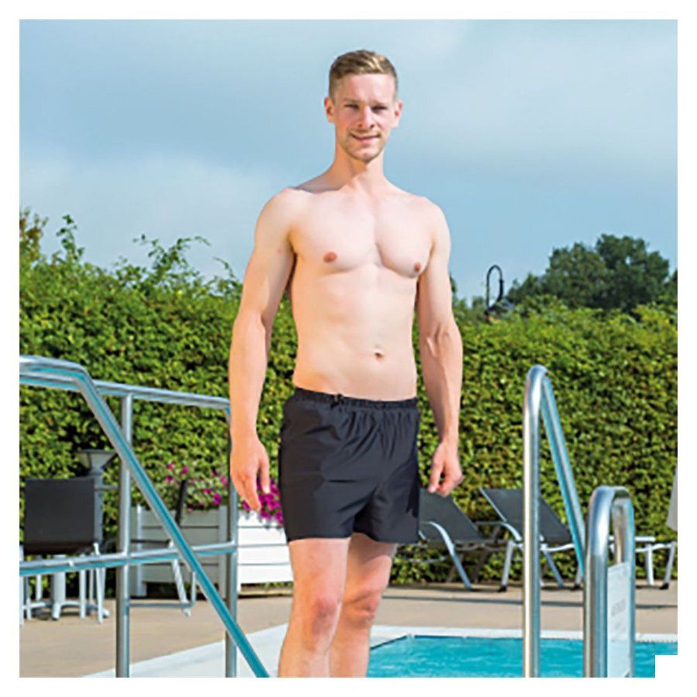 Homme en short de bain noir au bord d'une piscine. Regard vers la caméra. Arrière-plan : verdure et rambarde.