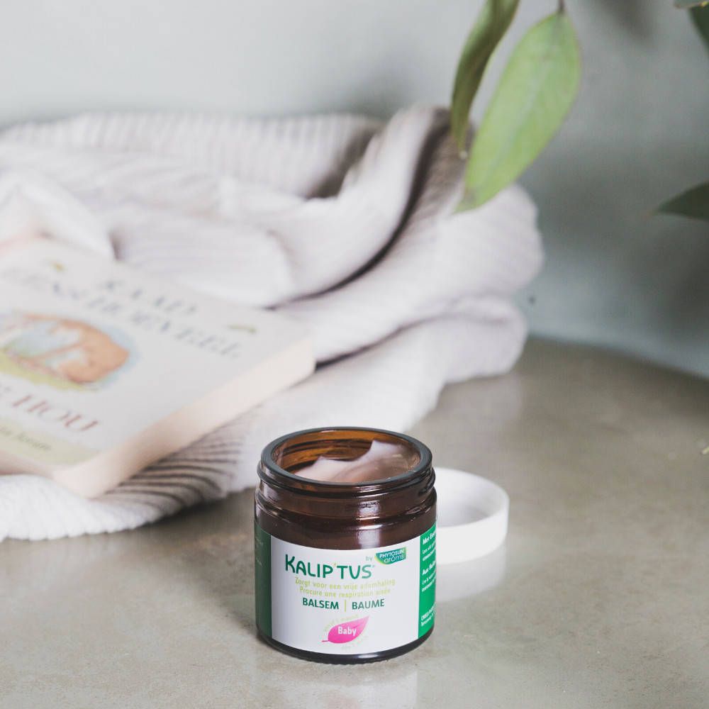 Bruine pot met witte deksel. Opschrift: Kalip'tus, Borstbalsem, Baby. Op een tafel.