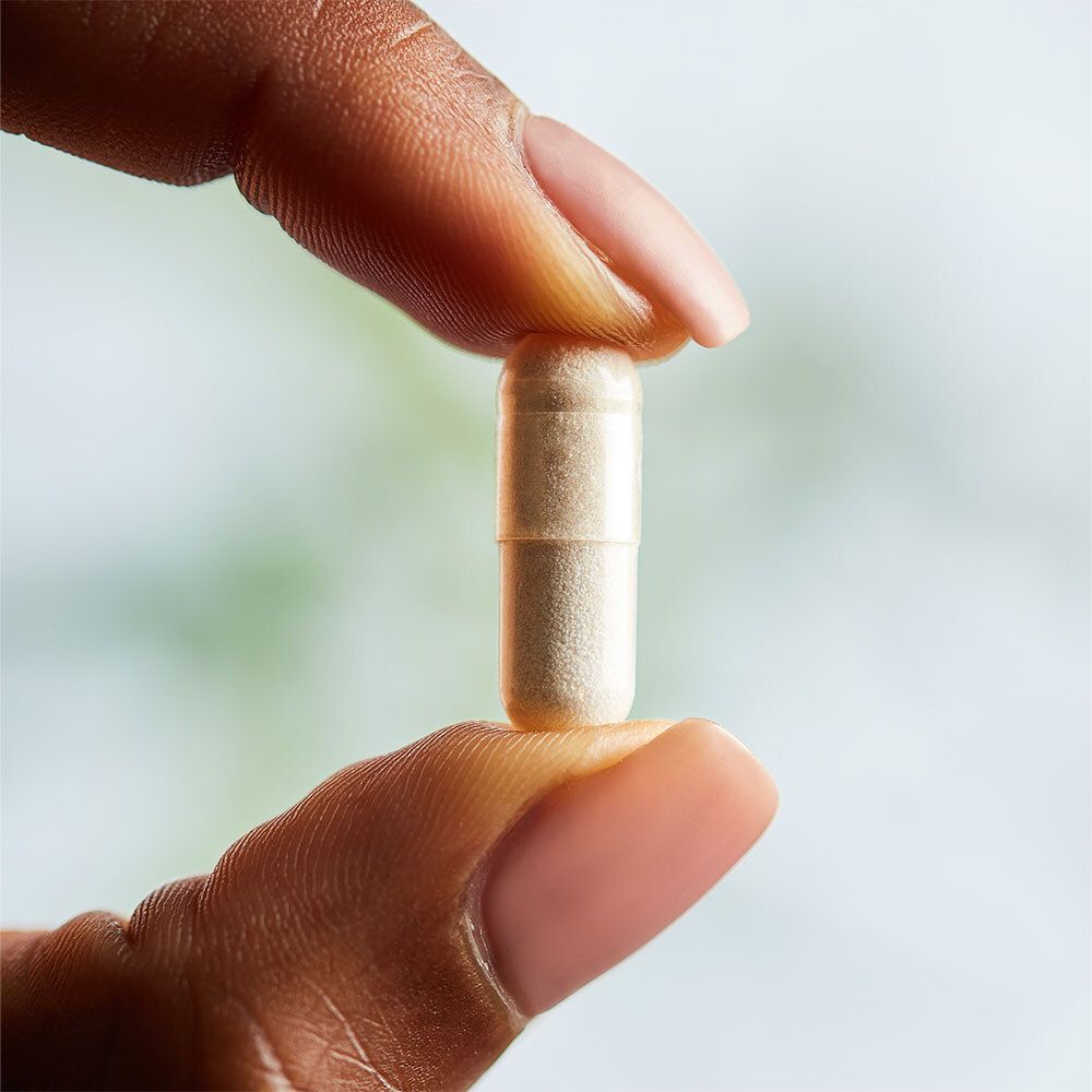 Une gélule tenue entre deux doigts. Couleur crème.