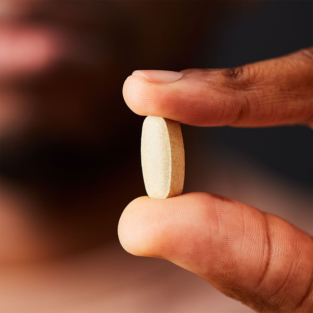 Personne tenant un comprimé entre le pouce et l'index. Le comprimé est ovale et beige.