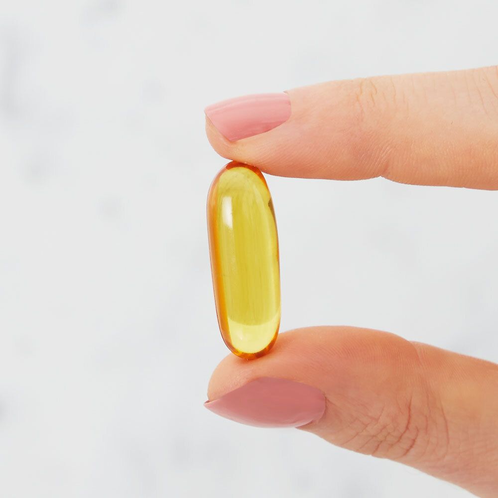 Gélule jaune ovale tenue entre deux doigts. Sur fond clair.