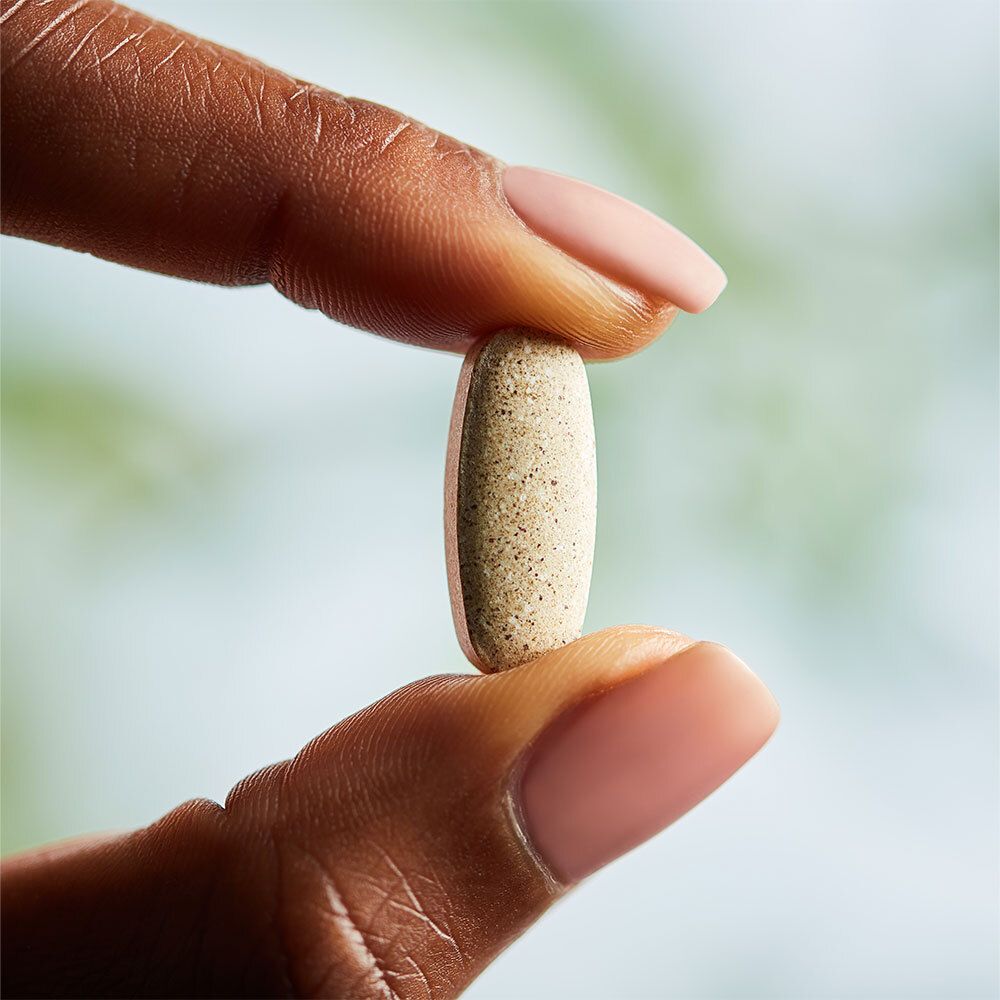 Un comprimé ovale beige est tenu entre deux doigts. Aucune inscription visible.