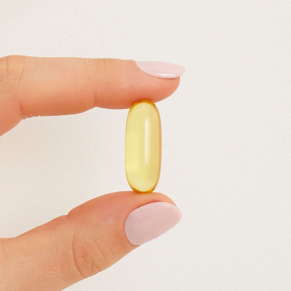 Een enkele, gele softgel capsule, vastgehouden tussen vingers. Duidelijke, ovale vorm.