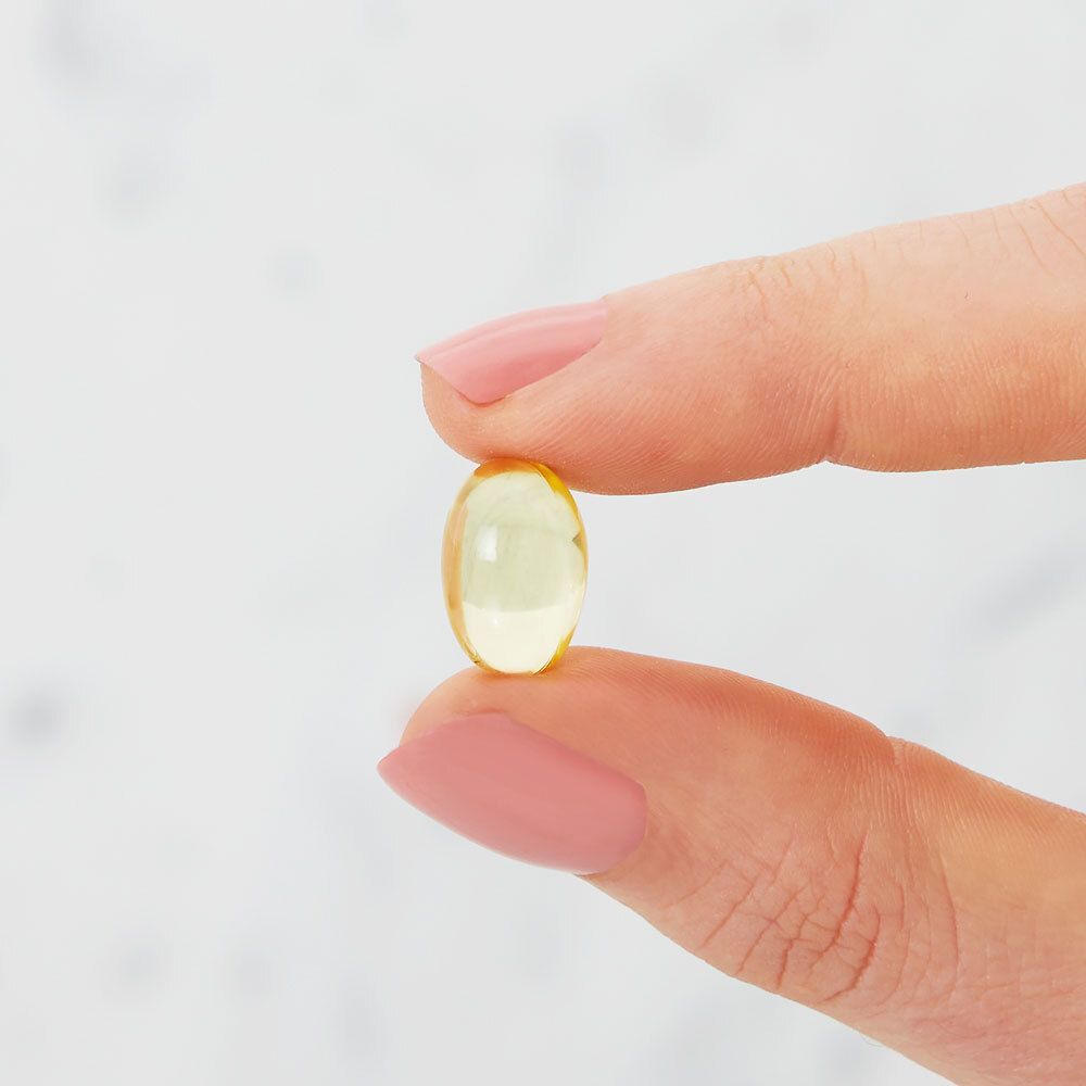 Close-up van een gele, ovale softgel capsule, vastgehouden tussen vingers. Lichte, vage achtergrond.