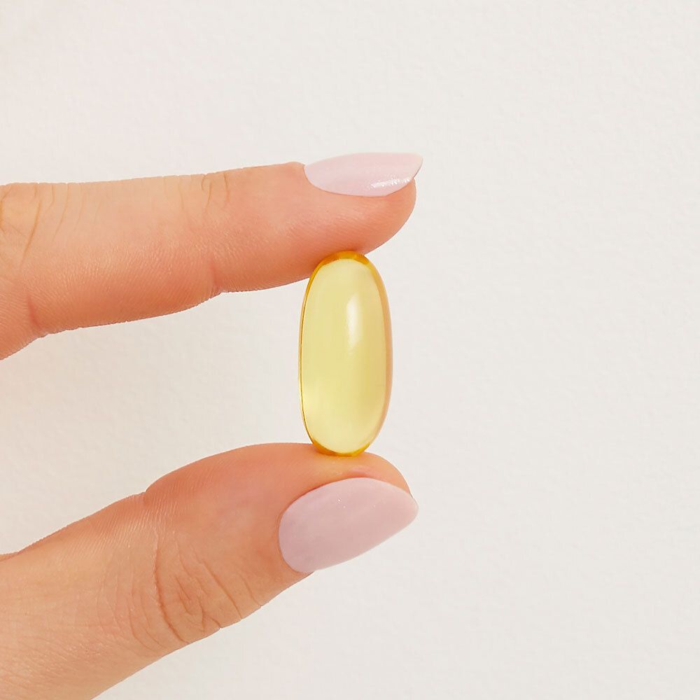 Main tenant une gélule ovale jaune. Ongles roses vernis.