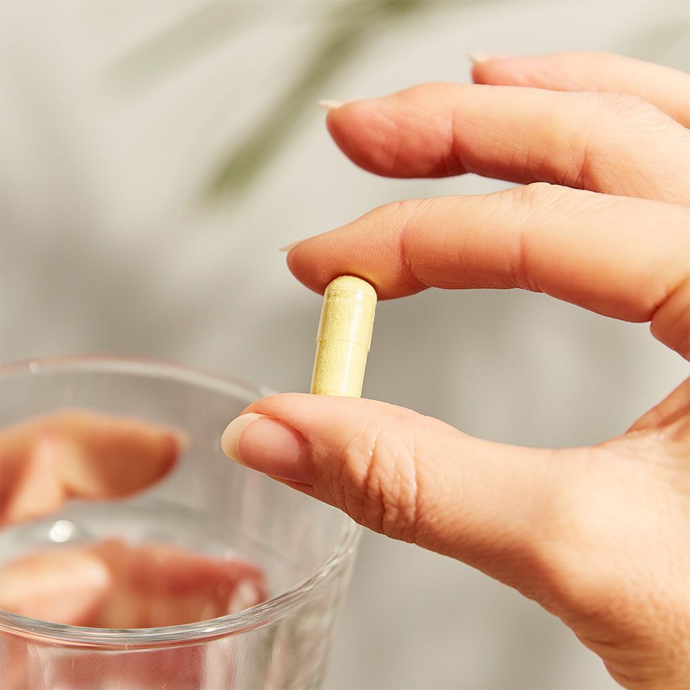 Main tenant une gélule jaune. Gélule dans la main, devant un verre d'eau.