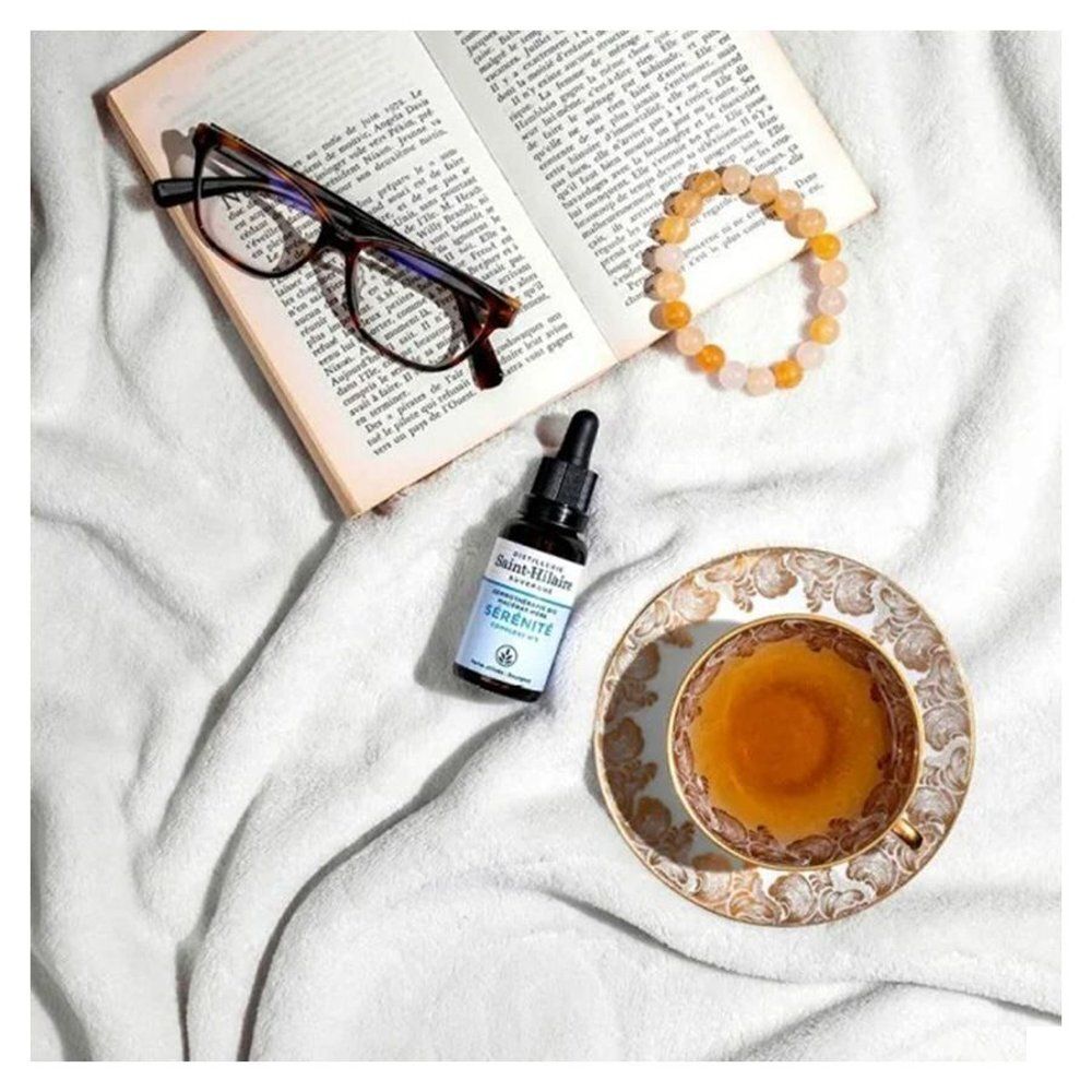 Flacon en verre brun avec compte-gouttes à côté d'un livre, lunettes, bracelet et tasse de thé sur tissu blanc.