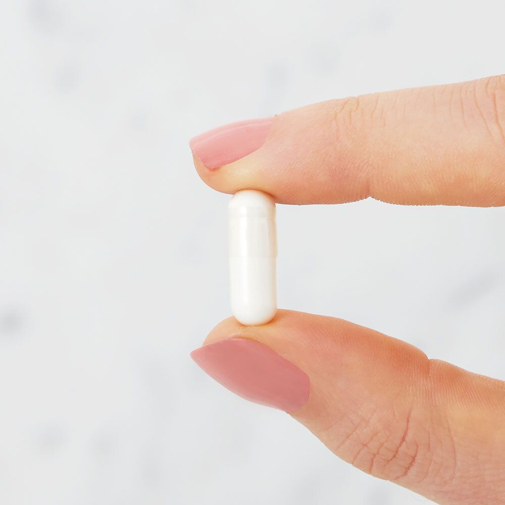 Une gélule blanche entre deux doigts. La gélule est oblongue et a une surface lisse.