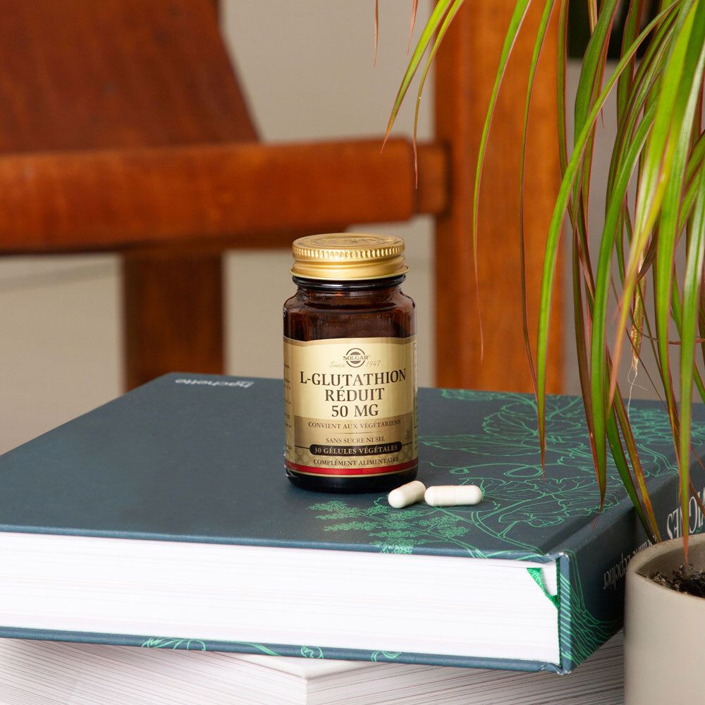 Le flacon est posé sur un livre. Deux gélules à côté. Une plante en arrière-plan.