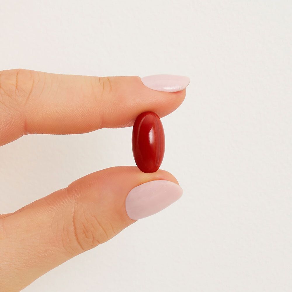 Une gélule rouge ovale, tenue par une main avec des ongles roses vernis.