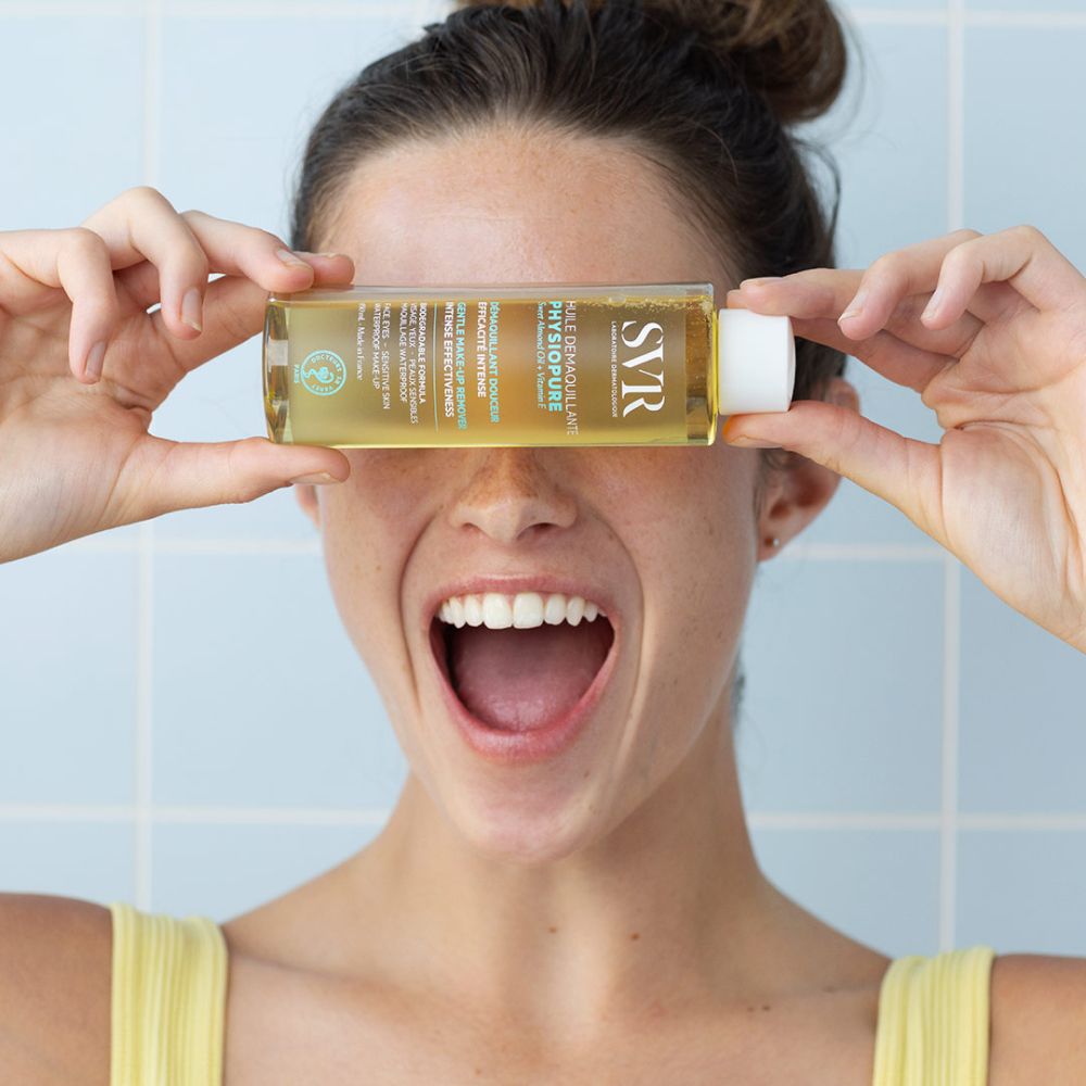 Femme tenant un flacon devant les yeux. Liquide jaune visible. Inscriptions SVR, PHYSIOPURE. Fond bleu.