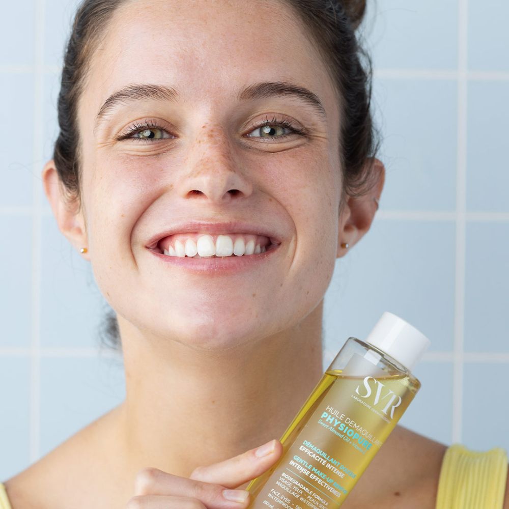 Femme tenant un flacon. Liquide jaune visible. Inscriptions SVR, PHYSIOPURE. Fond bleu. Femme souriante.