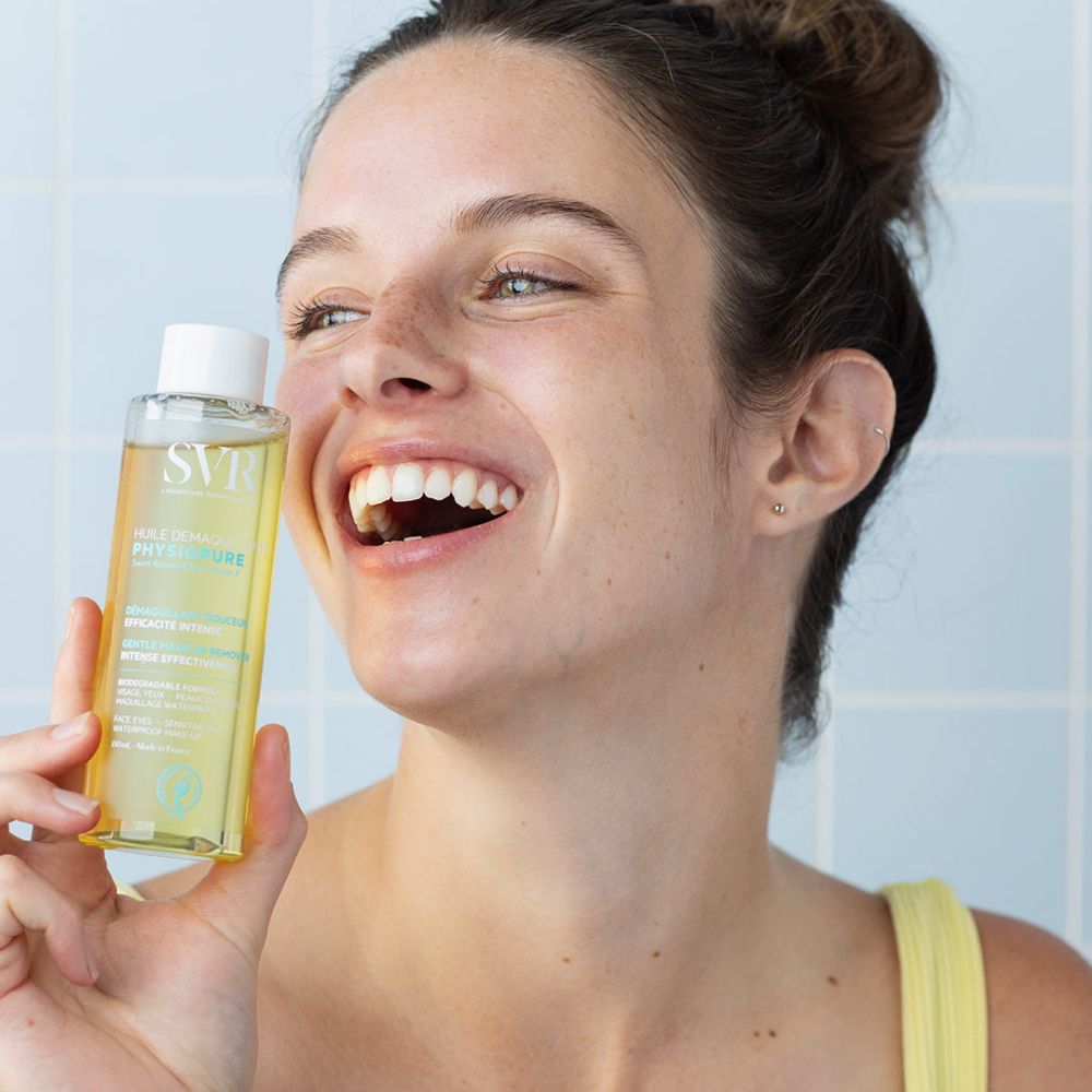 Femme tenant un flacon. Liquide jaune visible. Inscriptions SVR, PHYSIOPURE. Fond bleu. Femme souriante.