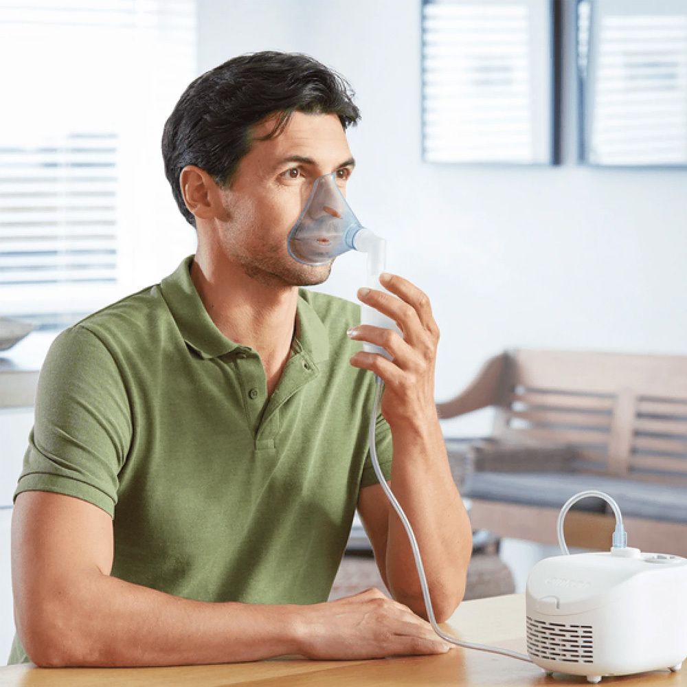 Man met masker verbonden met een witte compressor. Hij zit aan een tafel.