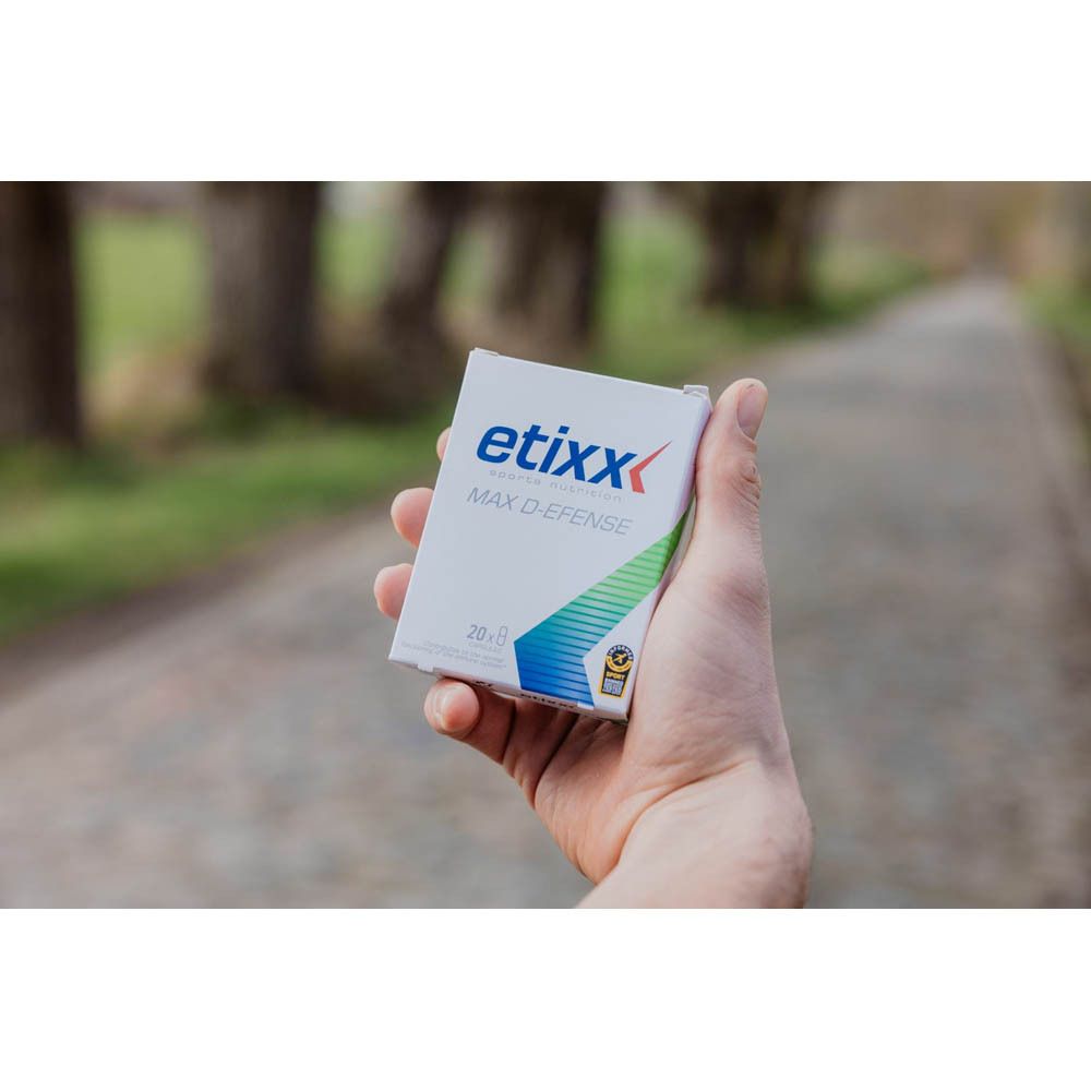 Main tenant une petite boîte blanche avec logo "etixx MAX D-EFENSE". Arrière-plan : chemin flou en extérieur.