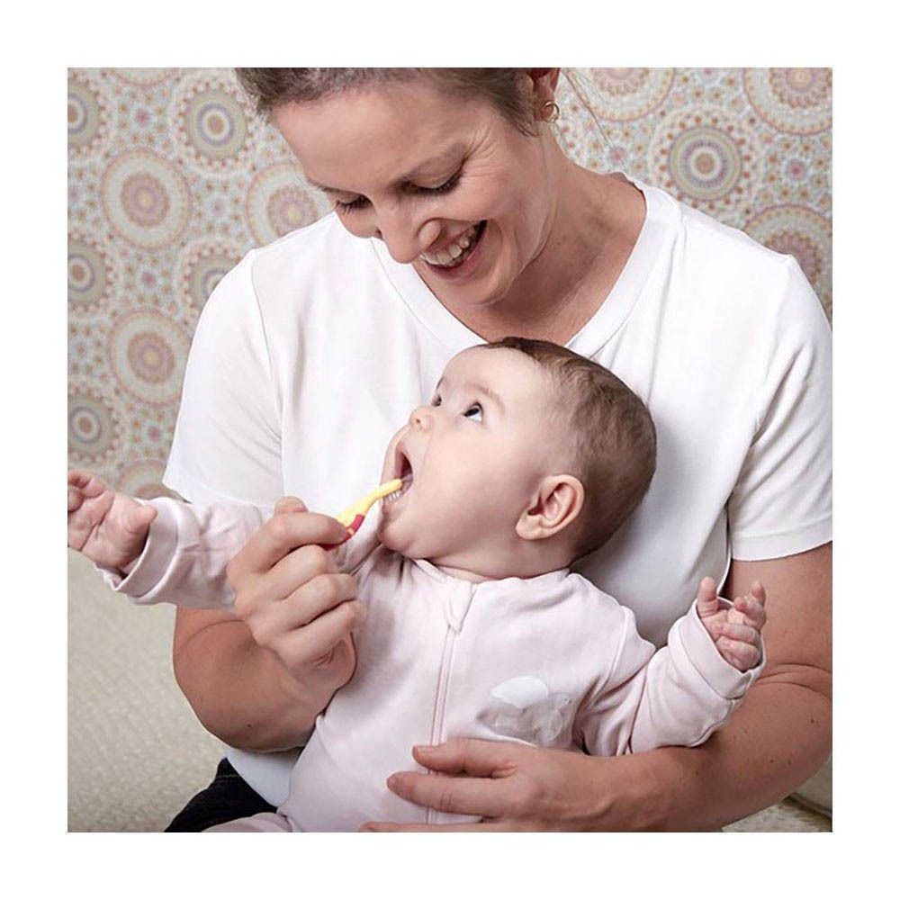 Baby met gele tandenborstel in de mond. Persoon houdt baby vast. Tandenborstel met roze handvat.