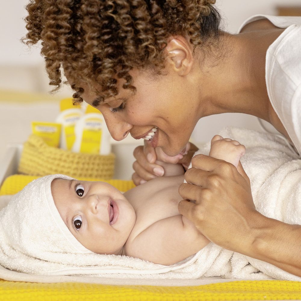 Une mère et un bébé. Le bébé est sur un tapis jaune, enveloppé dans une serviette. La mère sourit.