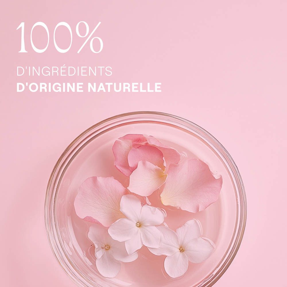 Pétales et fleurs dans un bol en verre. Fond rose. Texte: 100% d'ingrédients d'origine naturelle.