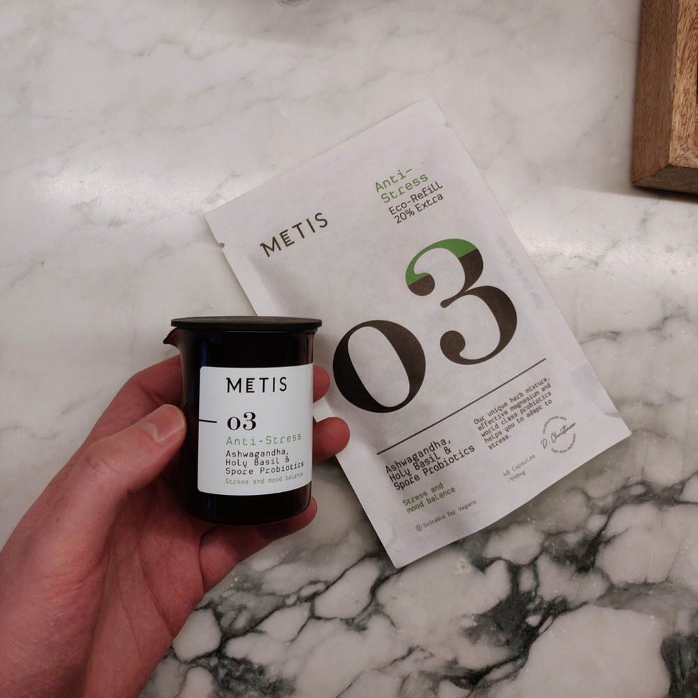Main tenant un pot noir et un sachet blanc. Pot avec logo METIS et "O3". Sachet : Anti-Stress, Eco-Refill, 20% Extra.