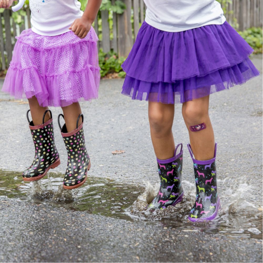 Deux enfants en bottes sautent dans une flaque d'eau. Un pansement est visible sur une jambe.