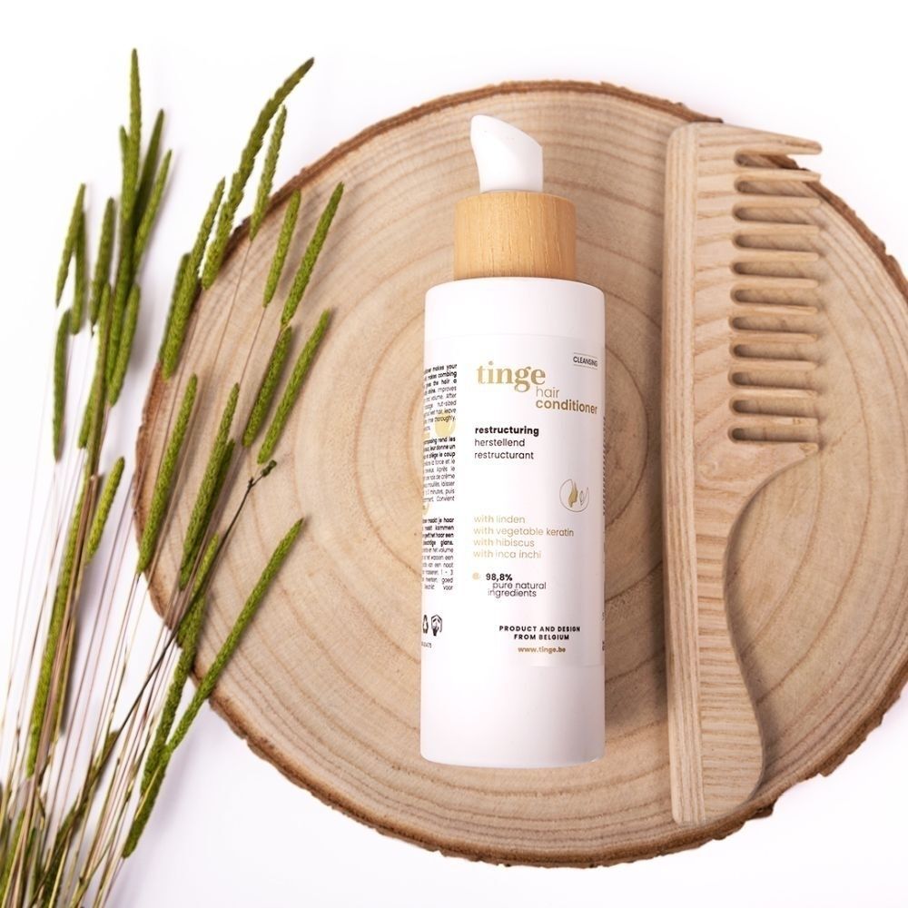 Flacon d'après-shampoing Tinge avec pompe et bouchon en bois, à côté d'un peigne en bois sur une bûche.