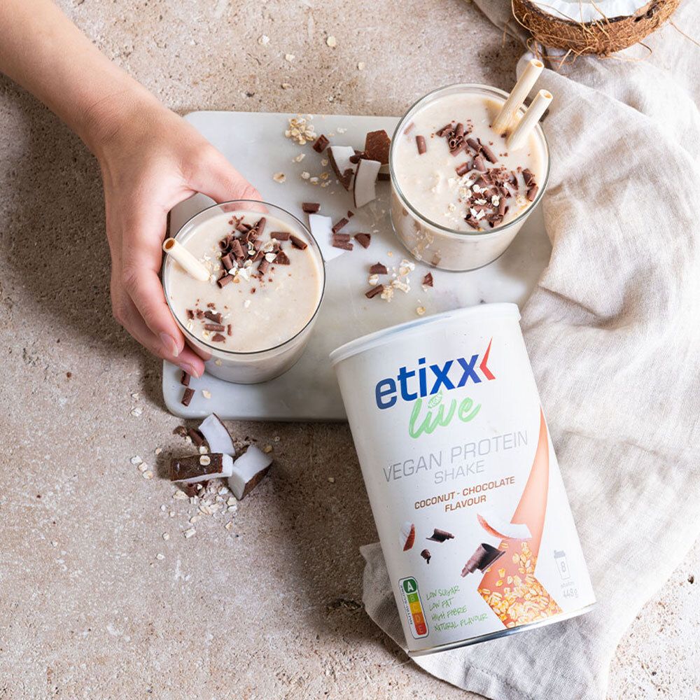 Twee glazen etixx Live Vegan Protein Shake. Kokos- en chocoladesnippers. Blik ernaast. Hand houdt glas vast.