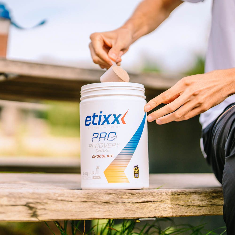 Persoon giet poeder uit een pot. Opschrift: etixx, PRO Line, Recovery Shake, Chocolate. Op houten bank.