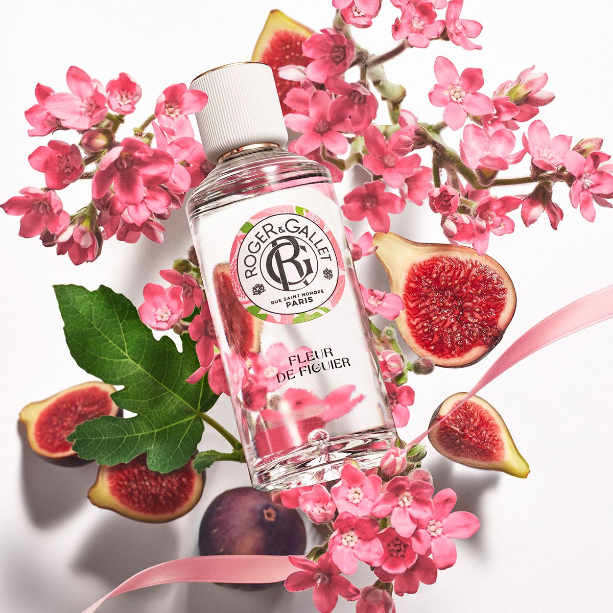 Flacon de Roger & Gallet Fleur de Figuier, entouré de fleurs roses et de figues. Flacon transparent avec bouchon blanc.