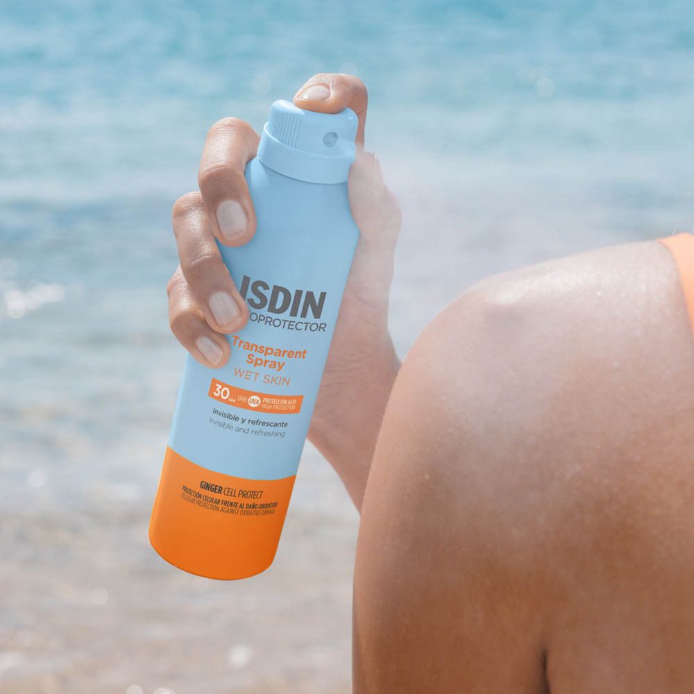 ISDIN Fotoprotector Transparent Spray SPF30. Flacon tenu à la main. Scène de plage.