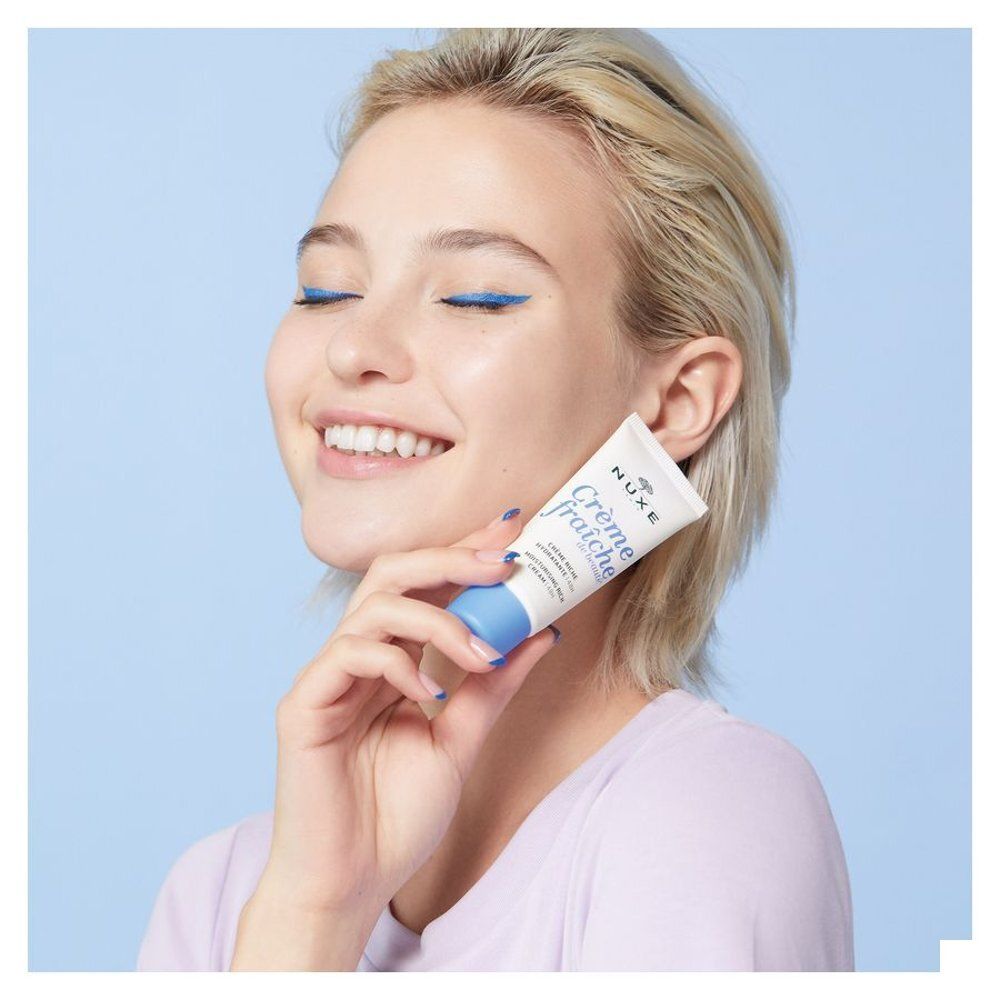 Vrouw houdt Nuxe Crème Fraîche de Beauté tube tegen haar wang, blauwe achtergrond.