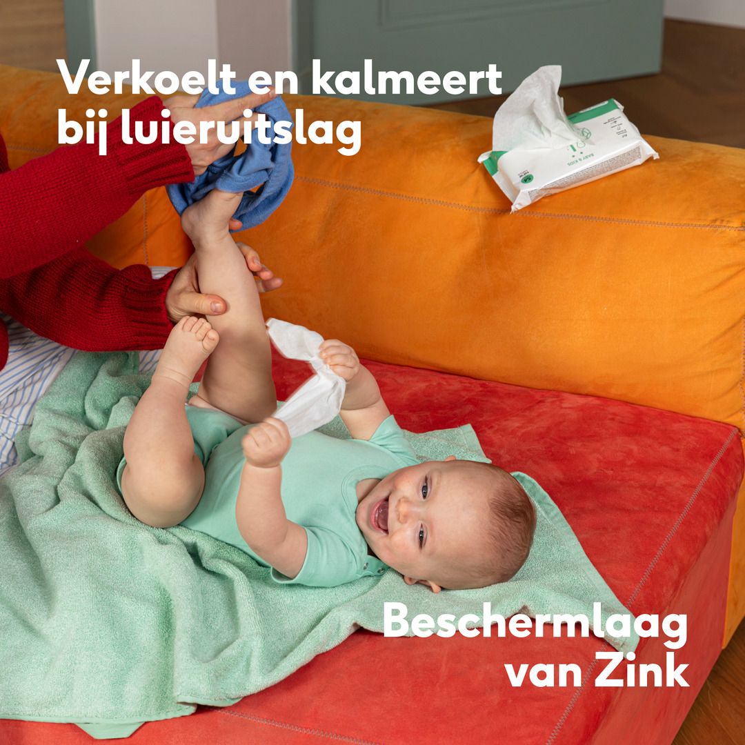 Baby wordt verschoond. Tekst: Verkoelt en kalmeert bij luieruitslag. Beschermlaag van Zink. Achtergrond: Oranje bank.