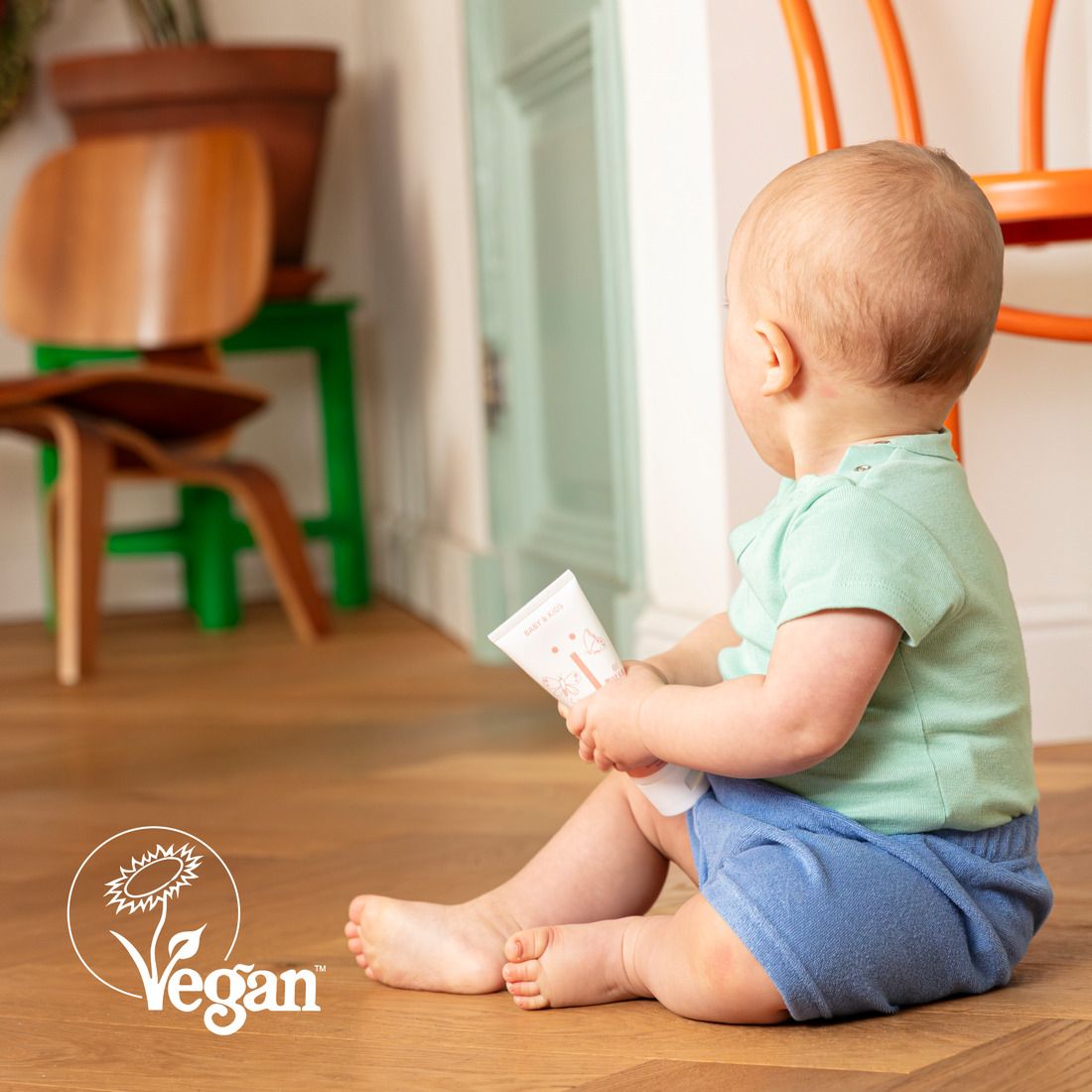 Baby zit op de grond met een tube. Vegan keurmerk. Achtergrond: Houten vloer, groene deur.