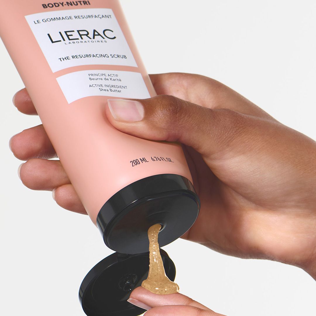 Main tenant un tube, la substance exfoliante sort. Tube rose avec bouchon noir. Texte: LIERAC, THE RESURFACING SCRUB.