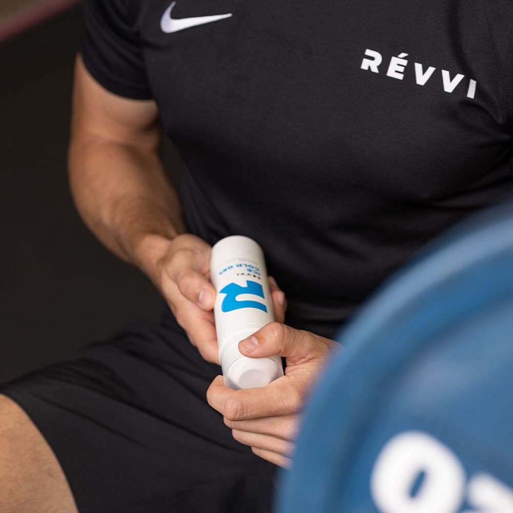 Persoon houdt een witte fles met blauw "R"-logo en tekst: RÉVVI ICE COLD GEL vast. Zwart T-shirt.