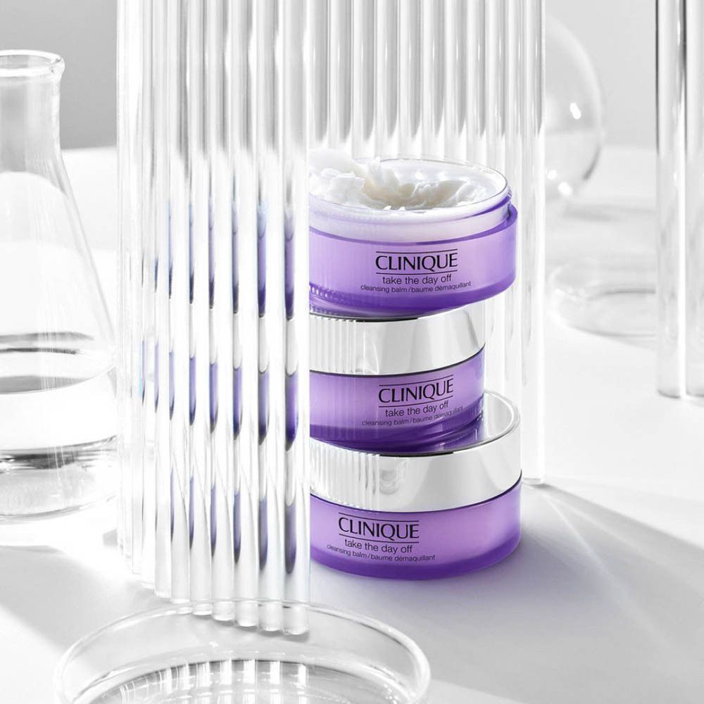 Drie potten CLINIQUE Take the Day Off, gestapeld. Glaswerk en laboratoriummateriaal op de achtergrond.