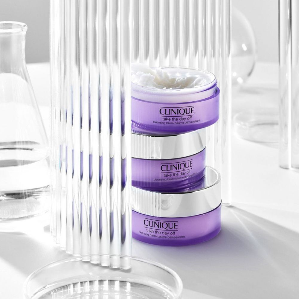 Drie potten CLINIQUE Take the Day Off, gestapeld. Glaswerk en laboratoriummateriaal op de achtergrond.