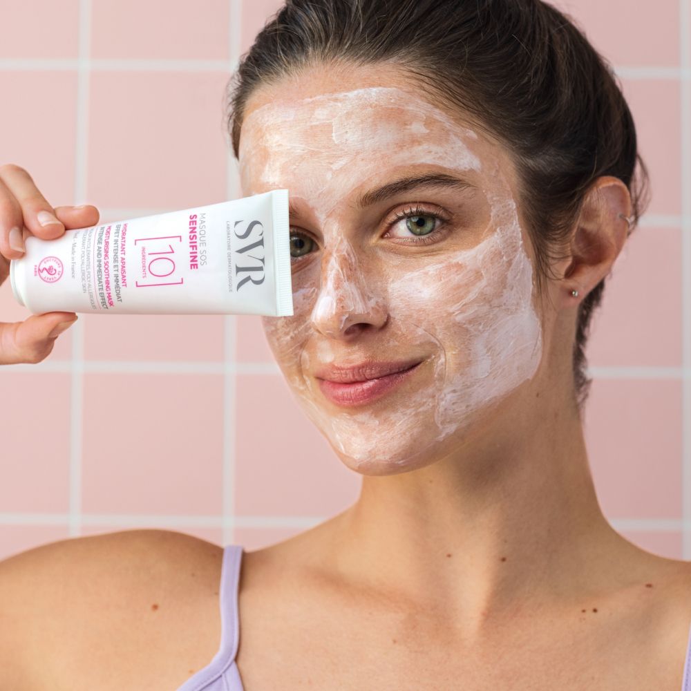 Femme tenant un tube devant son visage. Inscriptions: SVR, Masque SOS Sensifine, 10 Ingredients. Son visage est couvert de crème blanche.