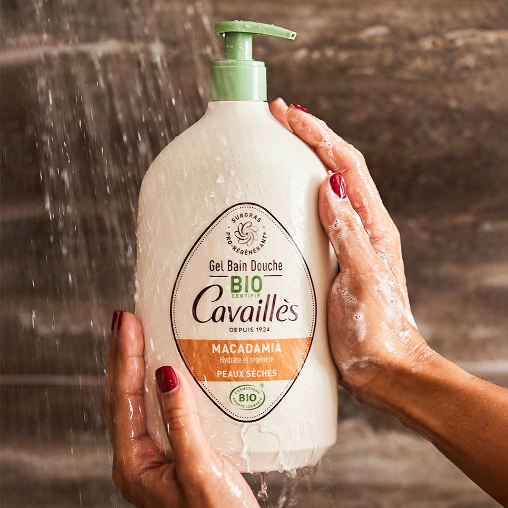 Handen houden een beige fles onder stromend water. Opschrift: Gel Bain Douche Bio Cavaillès, Macadamia. Rode nagels.