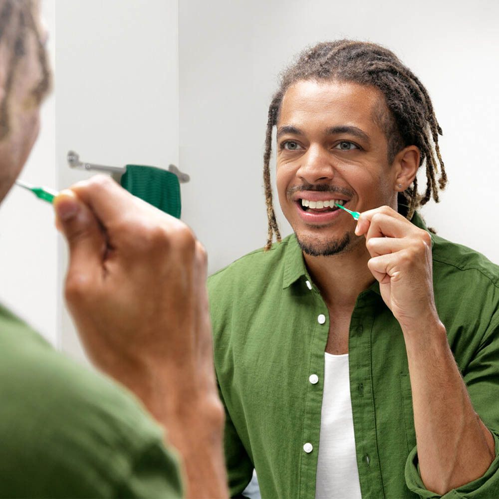 Un homme se brosse les dents avec un G.U.M Soft-Pick vert devant un miroir. Le pic est visible dans la bouche.