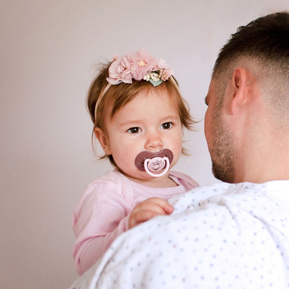Jeune enfant avec sucette en bouche, tenu par une personne. Bandeau rose.