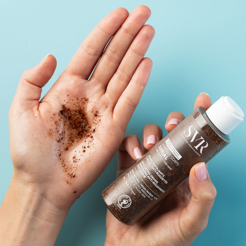 Handen met lichaamsscrub en fles. SVR-logo. XERIAL-tekst. Exfoliërende korrels.