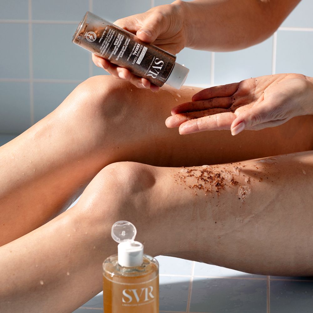 Persoon giet lichaamsscrub op hand en been. SVR-logo. XERIAL-tekst. Exfoliërende korrels.