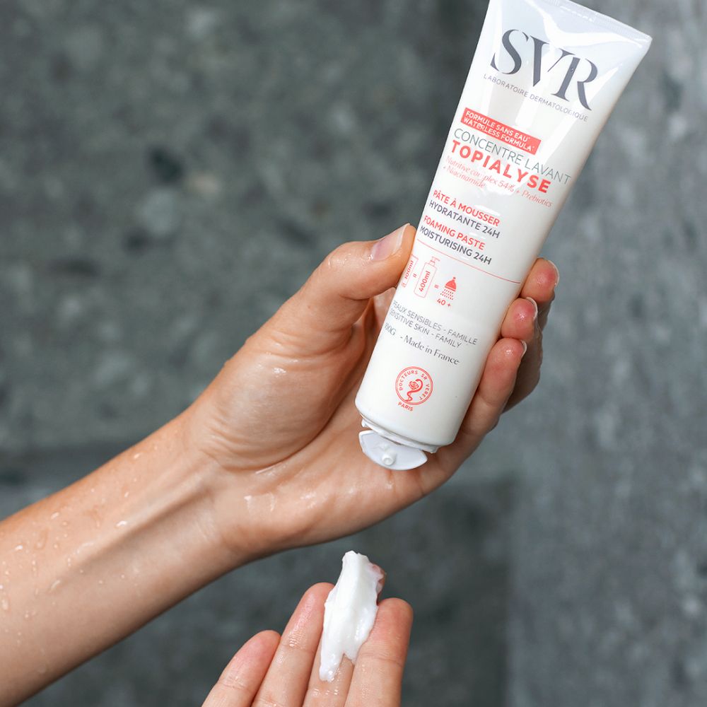 Hand houdt tube vast. Product wordt op hand aangebracht. Witte crème. Achtergrond: douche.