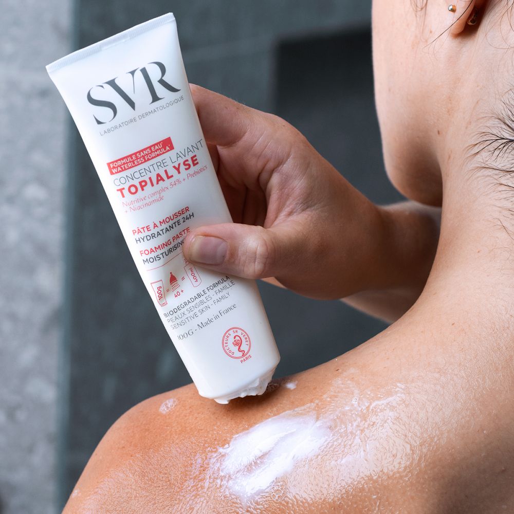 Persoon brengt product aan op schouder. Witte crème. Tube in de hand. Achtergrond: badkamer.