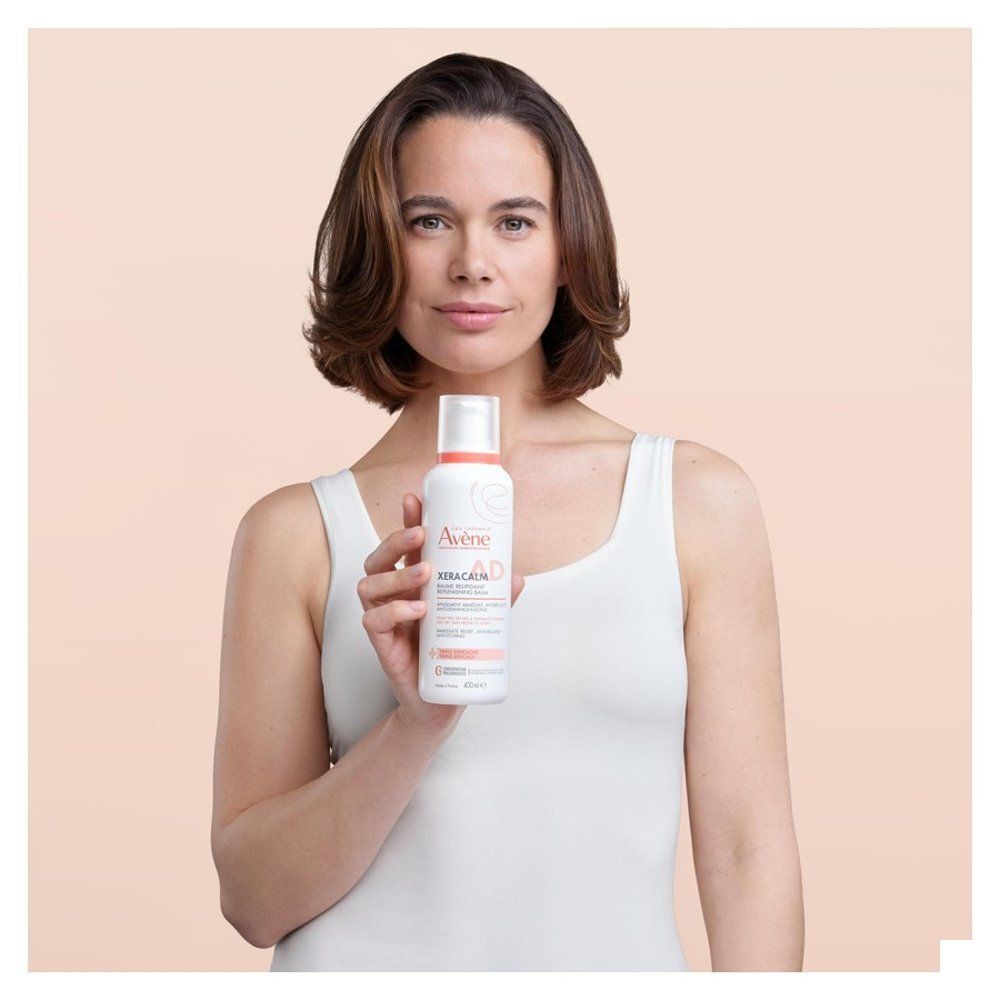Vrouw houdt fles vast. Opschrift: Eau Thermale Avène, XeraCalm A.D. Beige achtergrond.