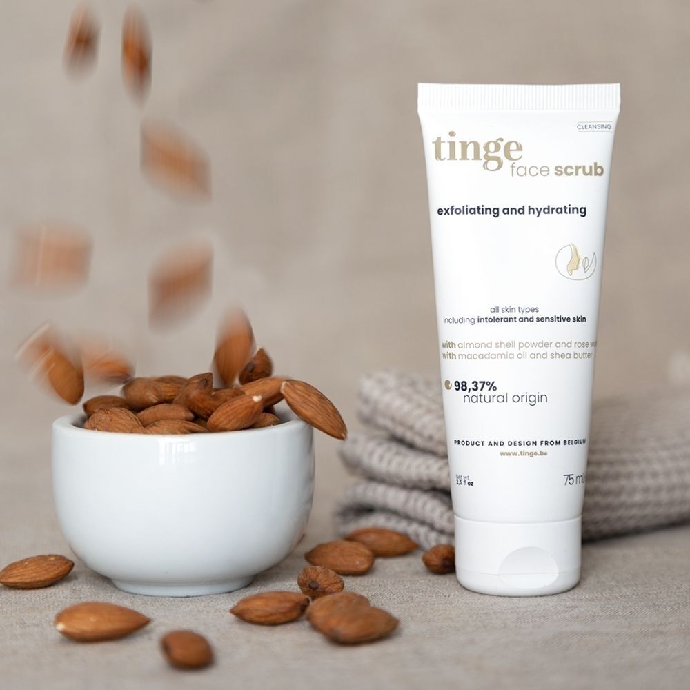 Tinge Gezichtspeeling tube met amandelen die in een kom vallen. 98,37% natuurlijke oorsprong.