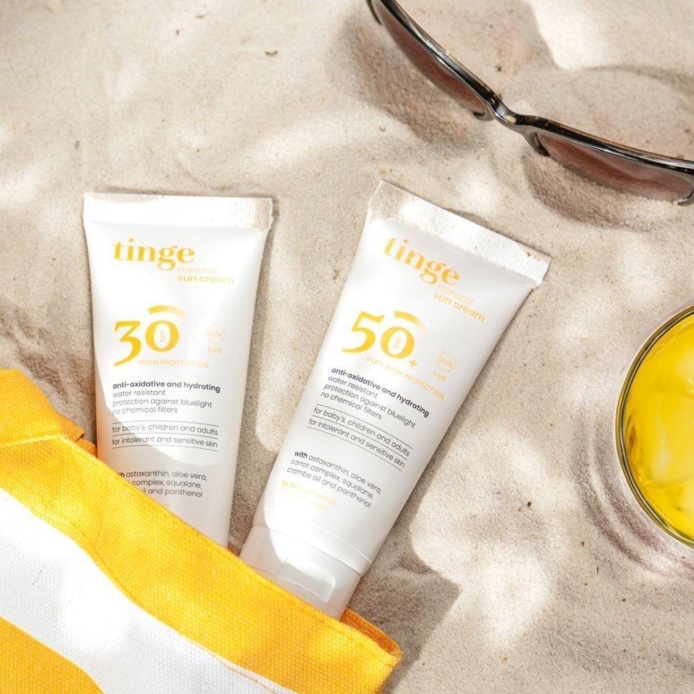 Twee Tinge zonnecrème tubes (SPF30 en SPF50+) op zand.