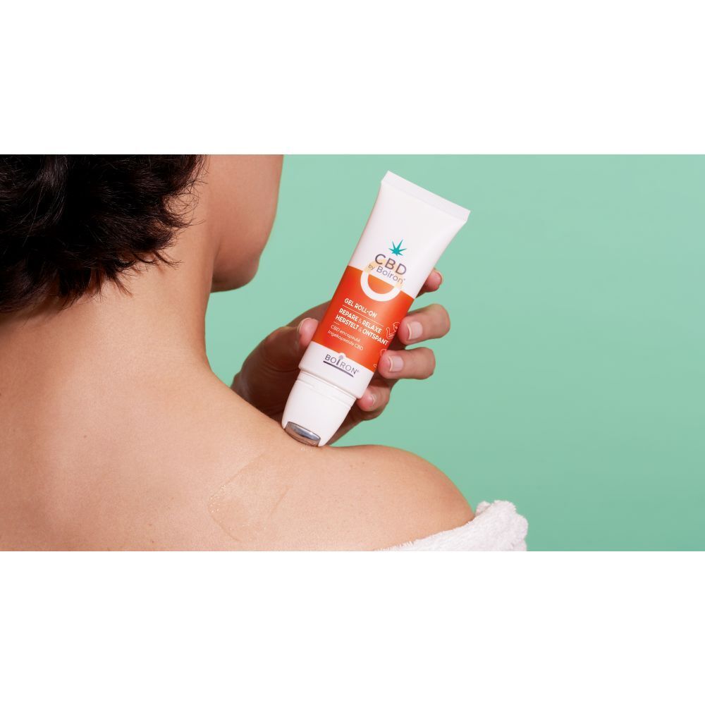 Persoon houdt CBD by Boiron gel roll-on vast. Product aangebracht op schouder. Groene achtergrond.